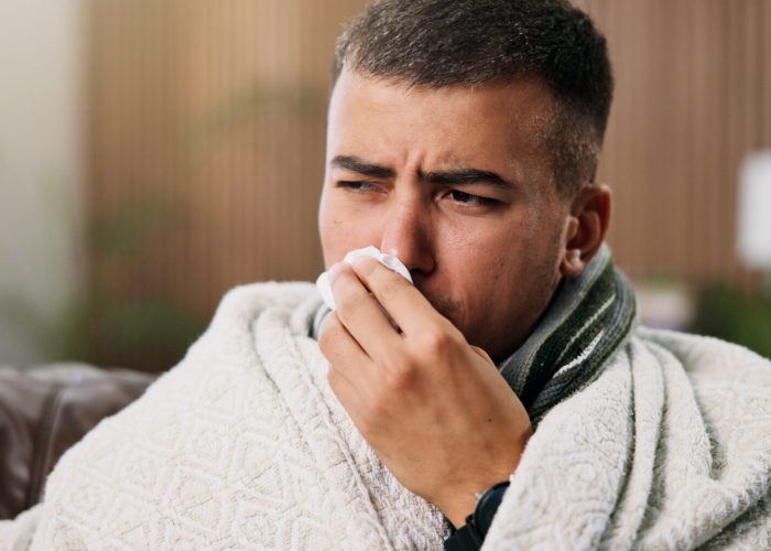 Man, tissue and blowing nose on sofa with blanket for comfort, flu and rest with recovery at home. Person, illness and cold symptoms with congestion, tired or allergies in living room at apartment.